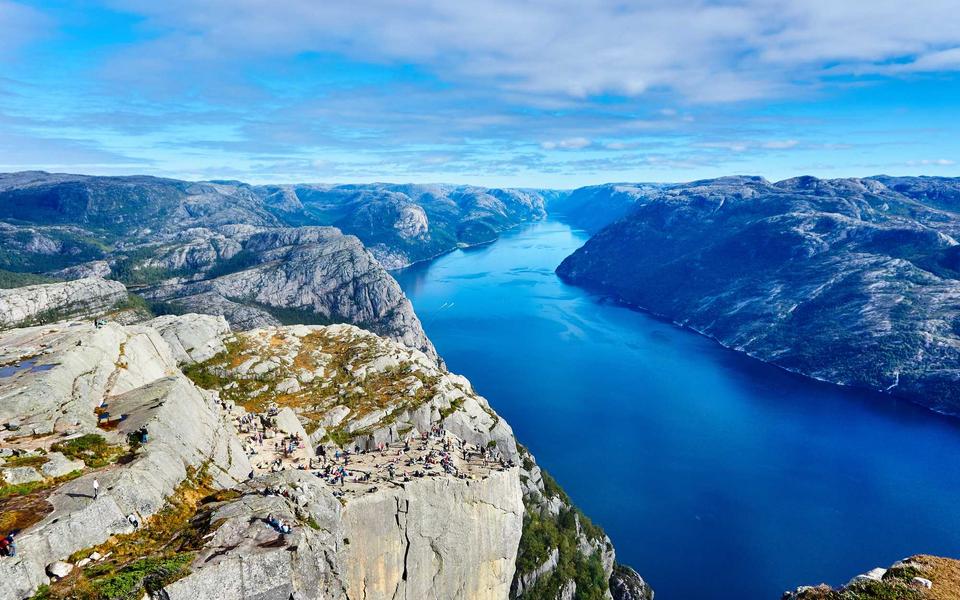A deep blue fjord between rocky cliffs, with hikers resting on a ledge in front